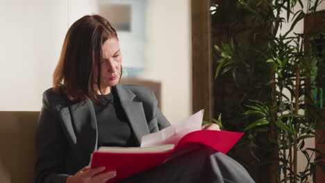 professional businesswoman in gray suit reading documents in red folder while seated indoors, appearing focused and serious, natural lighting with indoor plant and office decor in background
