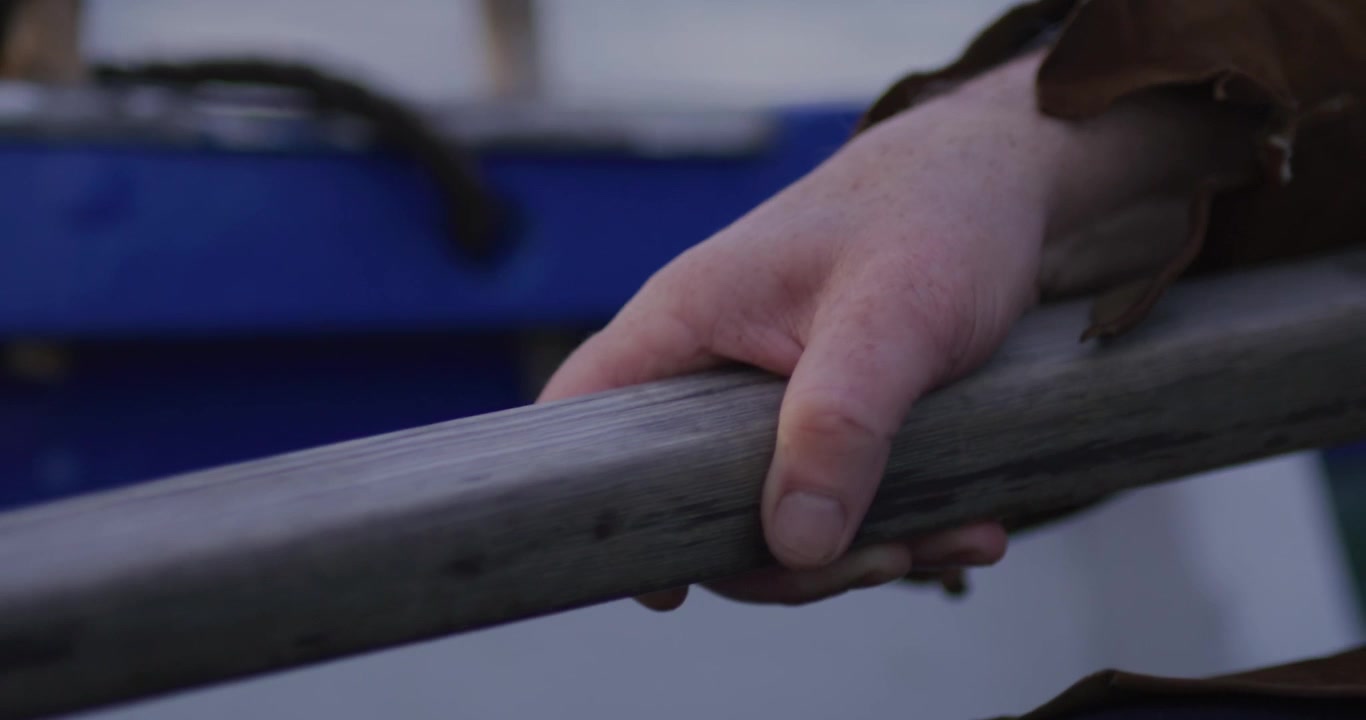 Close-up of a freckled hand gripping a weathered wooden bar, surrounded by leather and navy fabrics, with a dim, moody background suggesting effort, control, and action.
