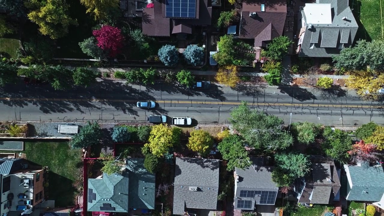 Ascesa aerea del drone sopra auto parcheggiate lungo una strada residenziale con asfalto crepato e fogliame autunnale