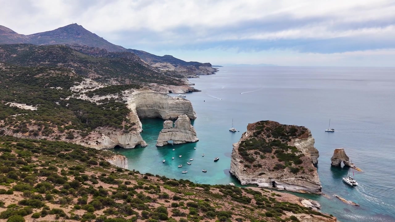 Et bredt luftfoto over Milos-kysten og Kleftiko-bugten, hvor blege havstakke, huler og ankrede både ligger under buskbevoksede bakker og et fjerntliggende bjerg under lagdelte skyer
