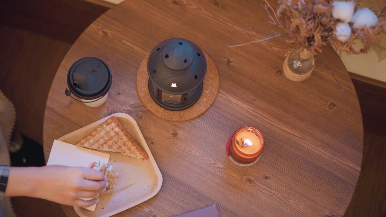 Fille attrapant un sandwich sur une table en bois avec café, bougie et objets décoratifs. La scène présente un éclairage doux provenant d'une lanterne allumée dans un intérieur aux tons chauds et à l'agencement cosy.