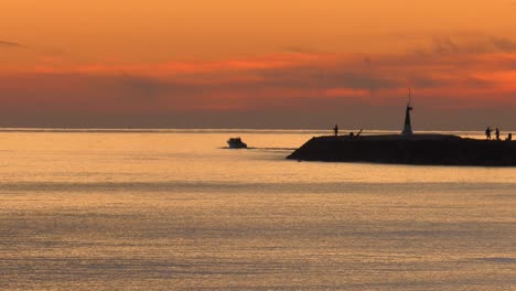 Lancha-A-Motor-Al-Amanecer-En-El-Mar-En-Calma-Pasando-Por-El-Muro-Del-Puerto-Con-Gente-Pescando