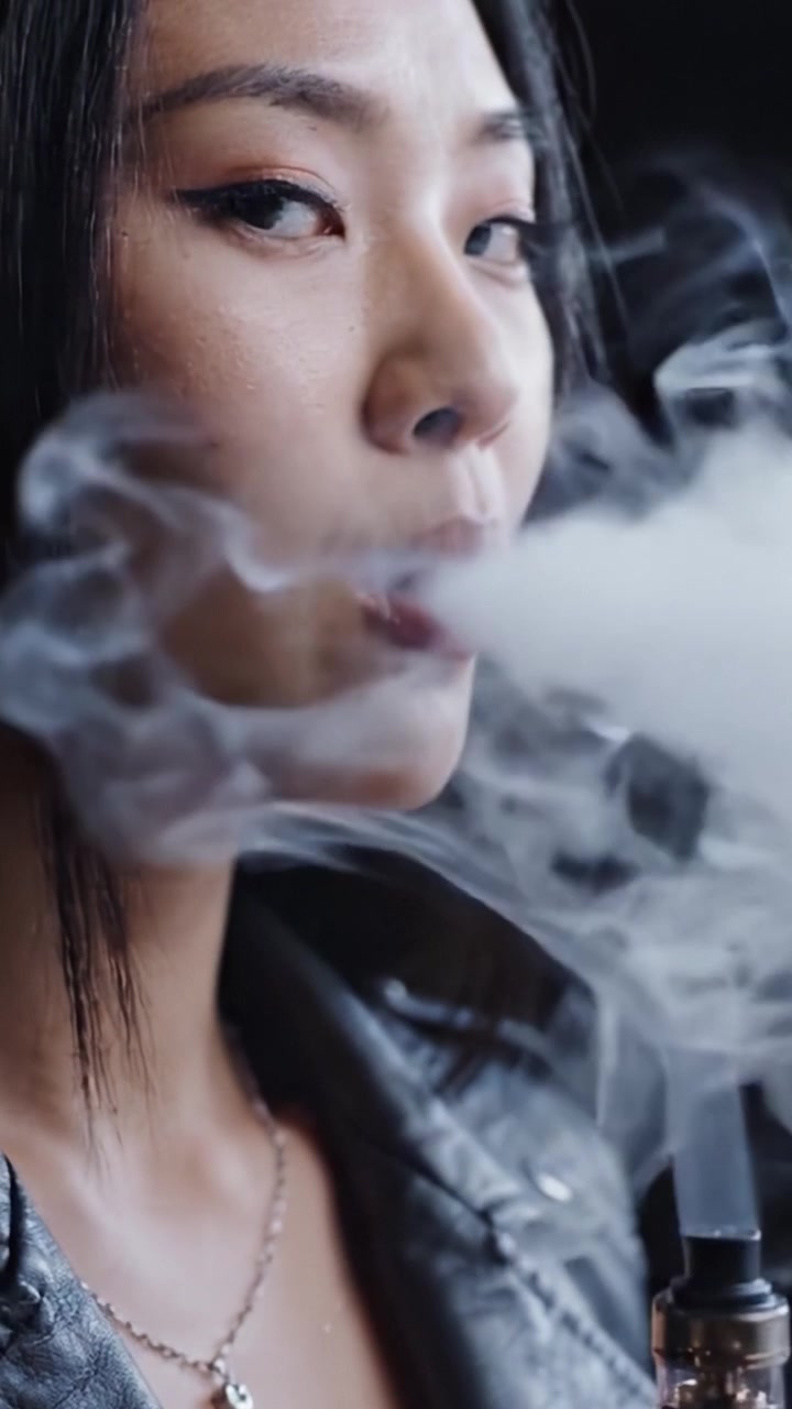 A Stylish Young Woman Embraces Vaping Culture, Exuding Confidence While Surrounded by Clouds of Vapor in a Dark, Atmospheric Setting