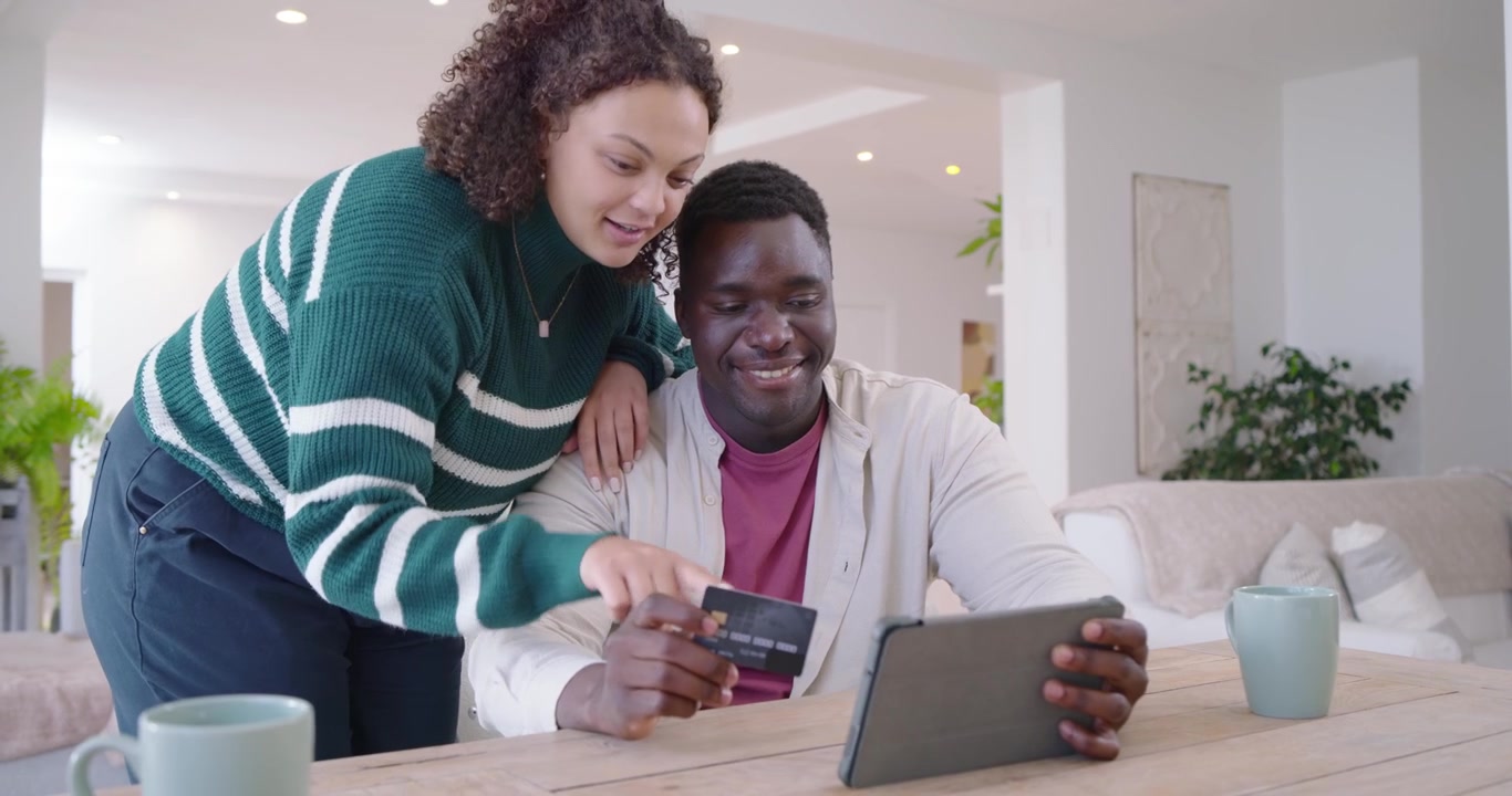 African American couple sitting at home, man holding card, woman guiding tablet to complete payment