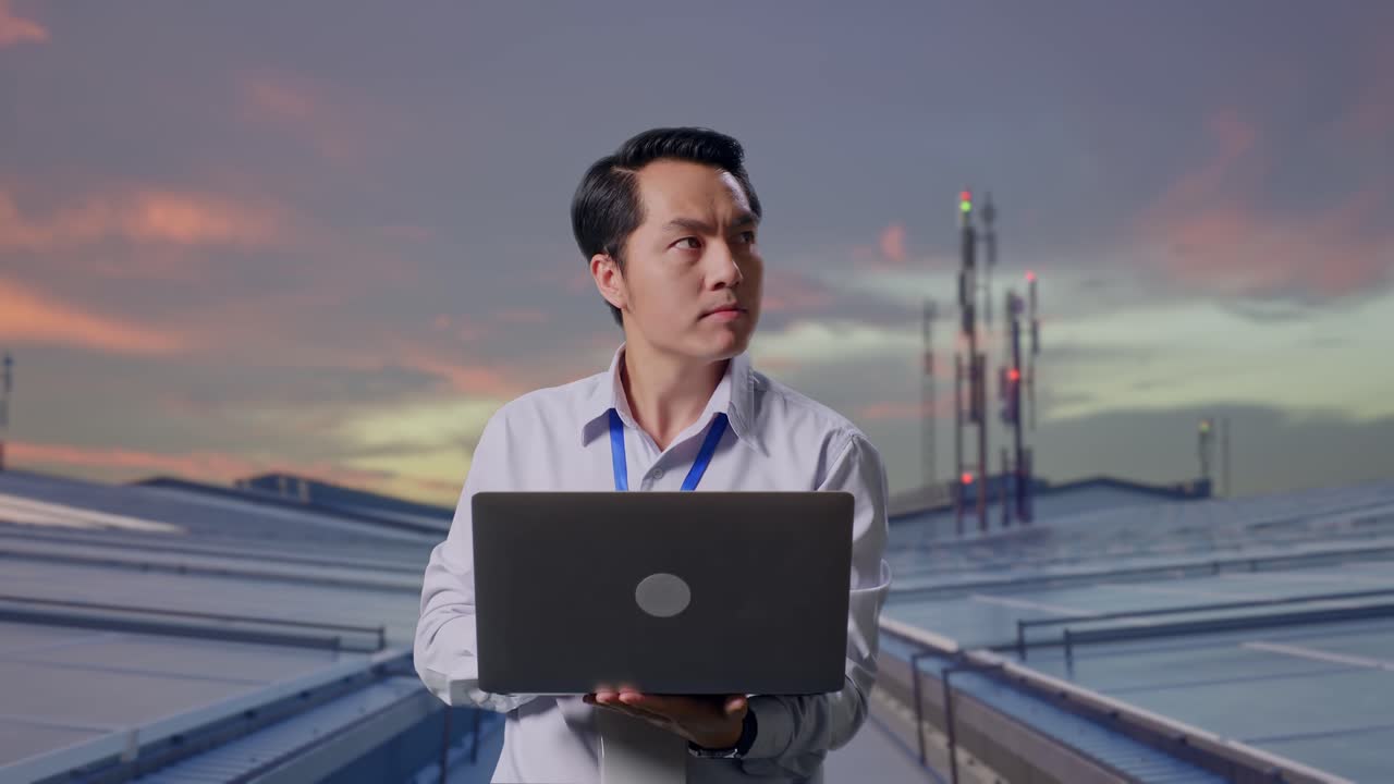 Aziatische mannelijke professionele werknemer staand met zijn laptop, inspecteert industriële zonnepanelen op het dak. Hij kijkt rond alvorens zich te concentreren op zijn laptop.