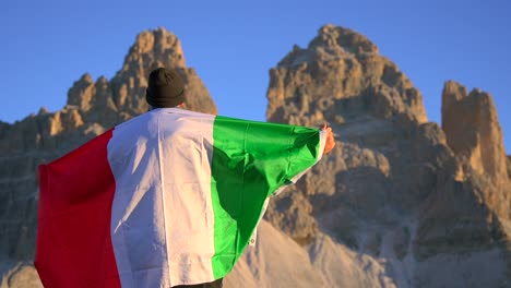 Imágenes-Filmadas-En-Rifugio-Auronzo,-Tri-Cine-En-Las-Montañas-De-Los-Dolomitas-Italianos-Con-Un-Hombre-Moviéndose-Y-Sosteniendo-Una-Bandera-Italiana-Y-Con-Hermosas-Montañas-En-El-Fondo,-Filmadas-Al-Amanecer-En-4k