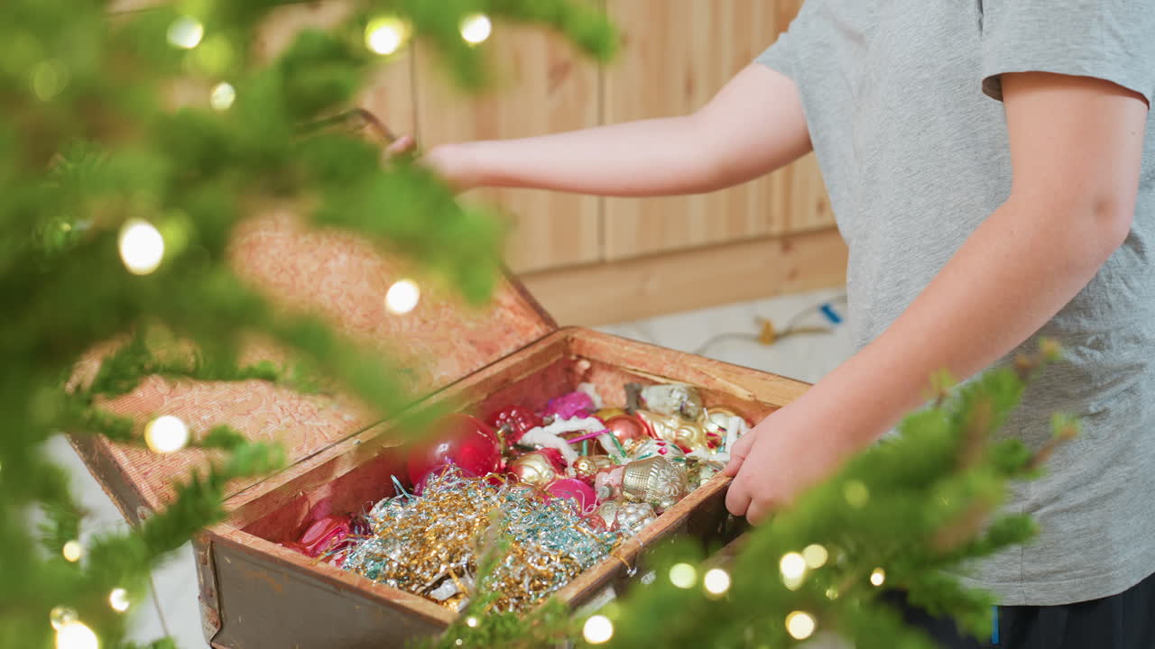 Partial view of young boy in casual grey shirt opening old box filled with colorful ornaments, carefully picking item to decorate christmas tree in cozy festive indoor setting with warm lights