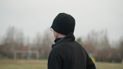 close-up of an individual wearing a black outfit and beanie, gazing towards a field, the background features a blurred view of people passing by