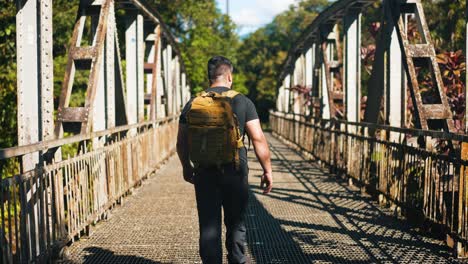 young man traveler with backpack walking on rusty old bridge outdoors on sunny summer day