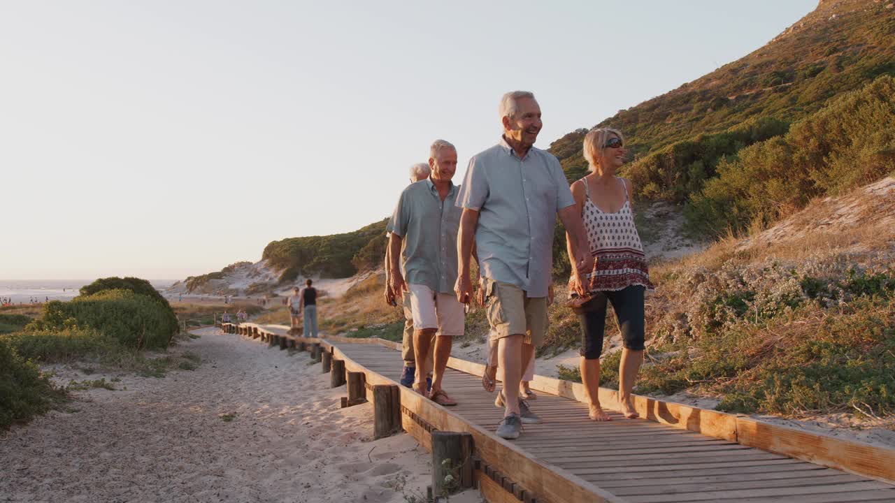 Group Of Senior Friends Walking Along Boardwalk At Beach On Summer Group VacationÊ