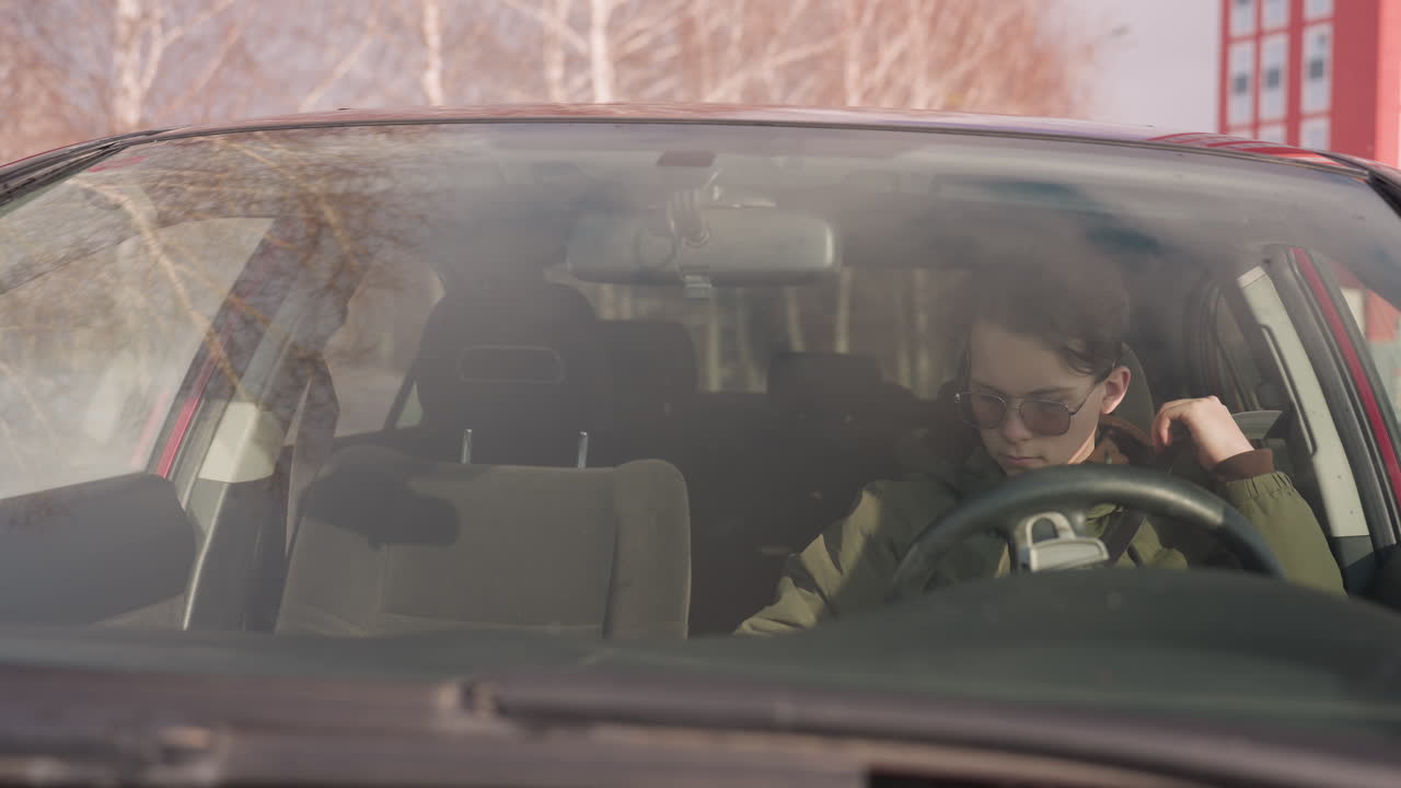 boy in parked car wearing sunglasses and winter jacket seen through windshield reaching for seatbelt with one hand while holding steering wheel with other, snowy outdoor background