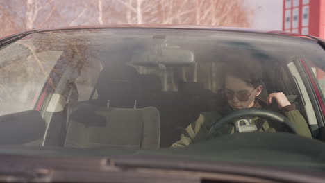 boy in parked car wearing sunglasses and winter jacket seen through windshield reaching for seatbelt with one hand while holding steering wheel with other, snowy outdoor background