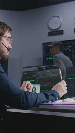 trader with pen in hand works at computer with displayed real-time stocks. colleagues analyze exchange market charts on big screens at background. office illuminated by blue light. vertical shot.