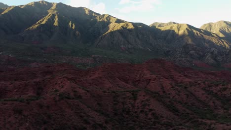 Barren-rolling-mountains-in-sun,-jagged-red-canyons-in-shadow,-aerial