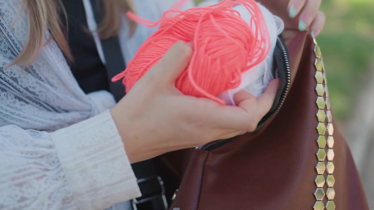 Primer plano de una mujer joven organizando hilo de colores en su bolso marrón con detalles hexagonales dorados. La escena muestra la textura y el patrón de la tela de encaje en la ropa, con iluminación suave en un entorno al aire libre.
