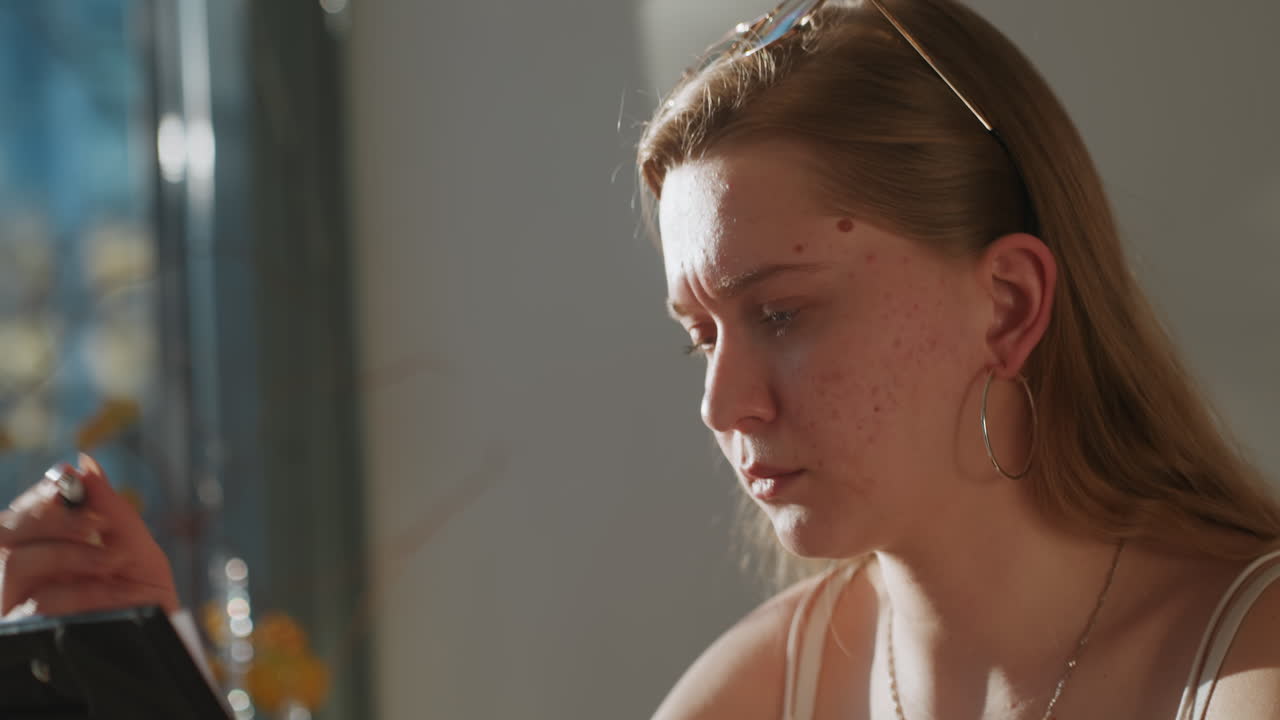 Young white woman reading tablet, tearful expression in warm window light, indoor kitchen table setting, hoop earrings and delicate necklace, focused downward gaze, emotional reaction to message, soft