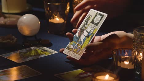 Close-Up-Of-Woman-Giving-Tarot-Card-Reading-On-Candlelit-Table-Holding-Death-Card-1