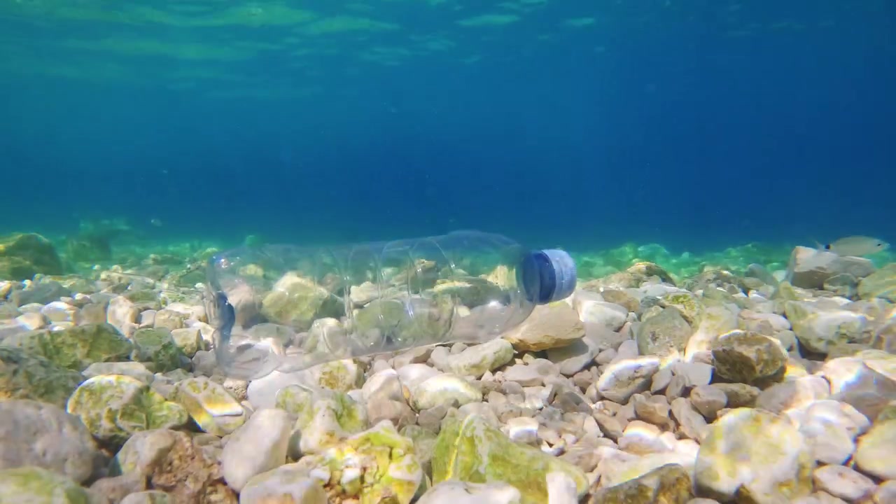 Uma garrafa plástica jaz no fundo do oceano, cercada por pedras e rochas. O ambiente subaquático mostra sinais de poluição plástica na água. Vida marinha se move nas proximidades.