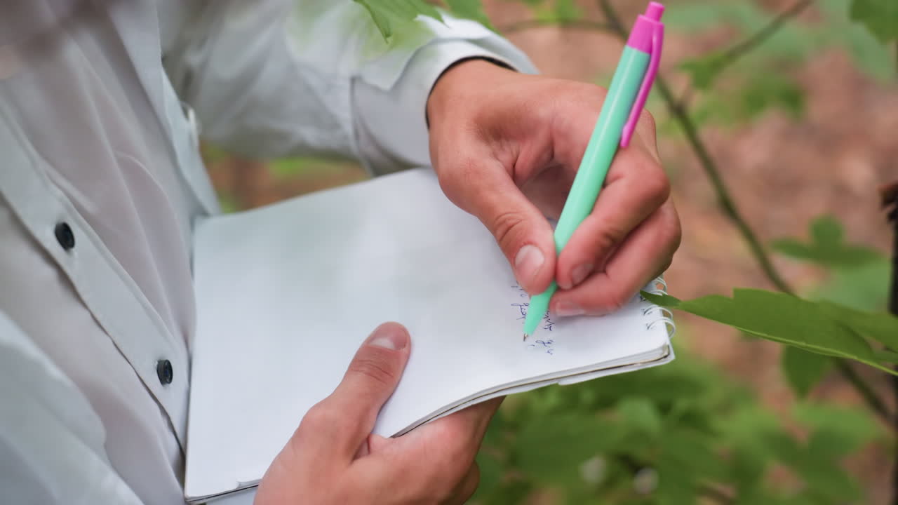 Hand view of researcher in white shirt writing notes on jotter with pen in forest, documenting ecological observations among green leaves and natural environment during scientific study