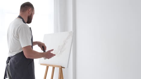 a young bearded artist of european appearance paints white paint on a clean white canvas - a young bearded artist of european appearance paints white paint on a clean white canvas. art studio in white tones