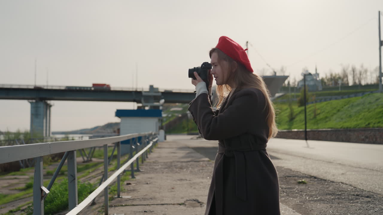 Beyaz tenli kadın köprü geçidini fotoğraflıyor, sokak fotoğrafçısı, bulutlu gökyüzü, kırmızı bere, uzun palto, kamera tutuyor, odaklanmış duruş, şehir nehir kenarı, korkuluklar, yansıtıcı su birikintileri, uzaktaki köprü.