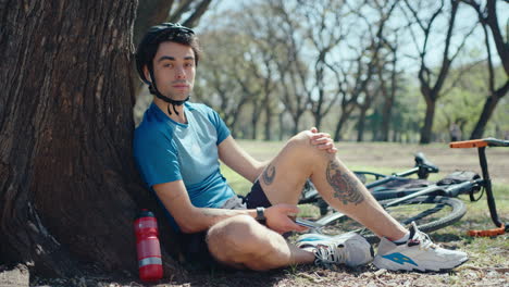 handsome cyclist using phone and posing on camera by tree