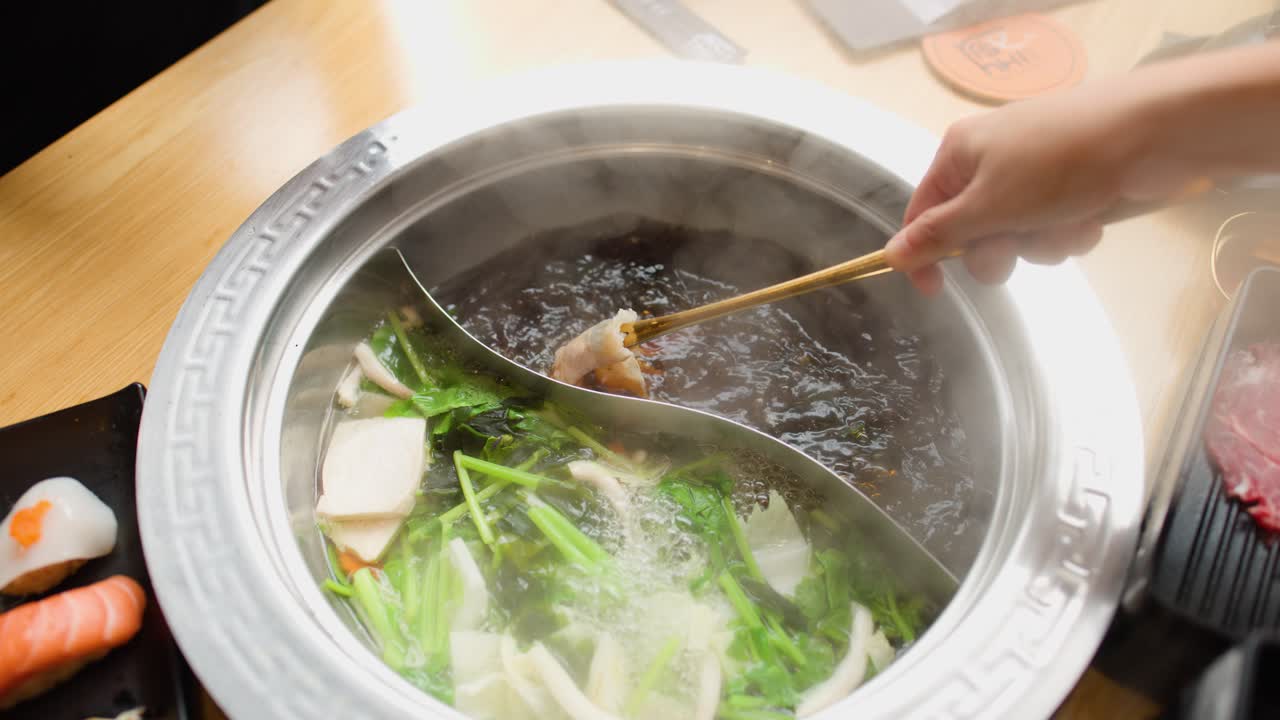 Hand cooks sliced meat in dual broth hotpot with fresh vegetables, overhead view, bright lighting