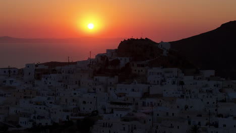 aerial drone telephoto trucking pan of anafi coastline with white homes at sunset, sun dipping below rugged cliffs, warm glow