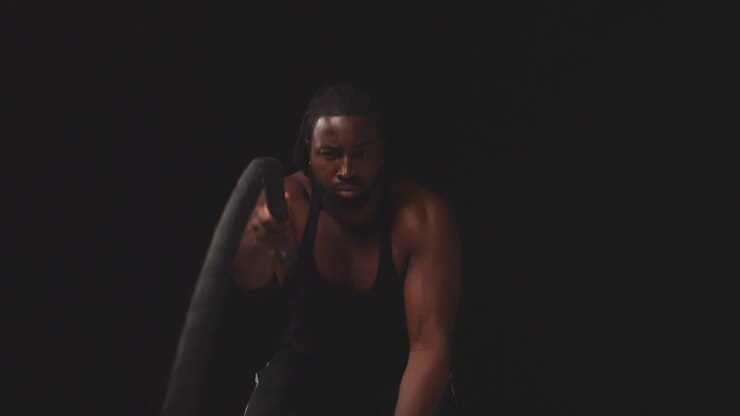 Studio Shot Of Male Athlete Training Wearing Vest Using Battle Ropes Shot Against Black Background