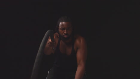 studio shot of male athlete training wearing vest using battle ropes shot against black background