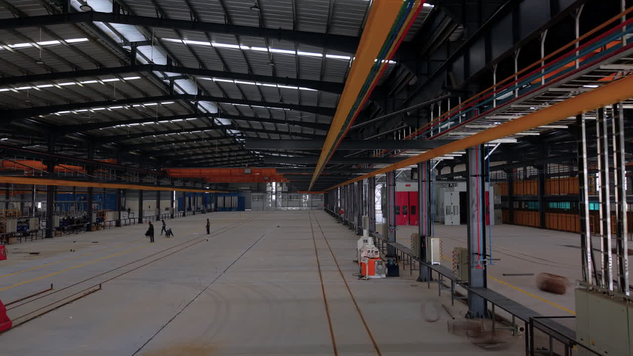 Bredt interiørbillede af et stort moderne lager- og produktionsanlæg med orange traverskraner, ingeniørudstyr og arbejdere. Perfekt til industriel fremstilling og logistik.
