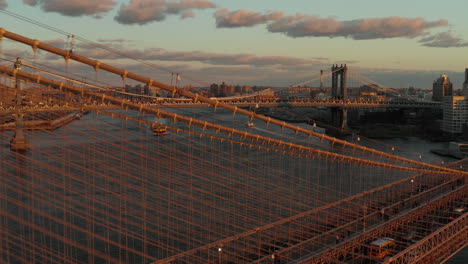 Fly-over-supporting-cables-of-Brooklyn-bridge.-Revealing-aerial-view-of-cargo-boat-passing-under-Manhattan-Bridge.-Sunset-in-city.-Brooklyn,-New-York-City,-USA