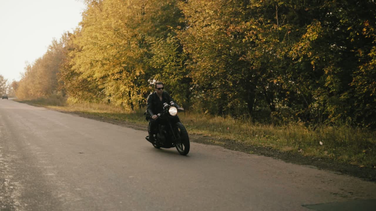joven elegante con gafas de sol y chaqueta de cuero montando una motocicleta en una carretera de asfalto en un día soleado en el bosque en otoño