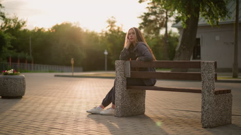 lady seated on bench outdoors during golden hour, resting her face on hand, with contemplative expression as sunlight softly illuminates her, surroundings include paved path, greenery and building