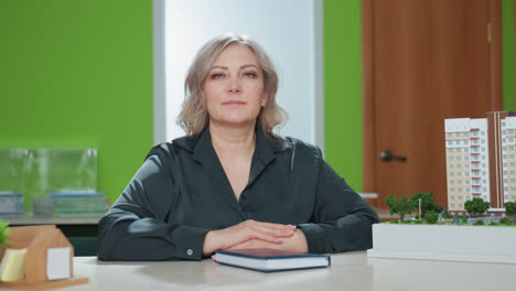 realtor in professional attire seated confidently at desk with hands resting on closed book, head tilted slightly and calm expression, positioned beside detailed architectural building model