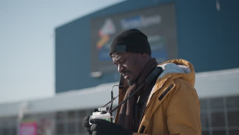 city dweller walking snowy street holding beverage cup and camera, glancing upward toward skyline as sunlight illuminates winter scene with parked cars and distant buildings under daylight shadows