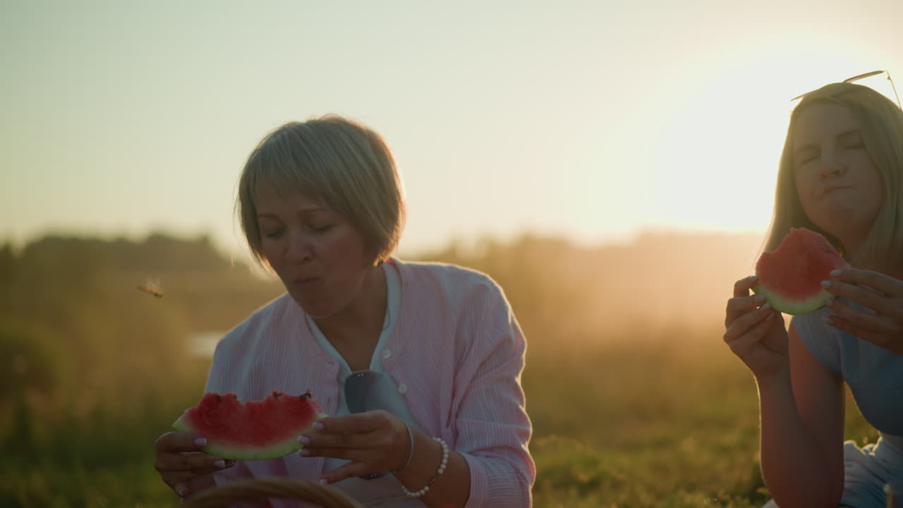 두 친구가 해가 지는 동안 야외에서 신선한 수박을 즐기고, 한 명이 잠시 멈추고, 근처에 떠다니는 용벌레에 매료되어, 빛이 그들의 미소를 비추고 있습니다.
