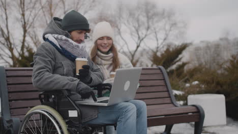 Hombre-Discapacitado-En-Silla-De-Ruedas-Y-Su-Amigo-Viendo-Algo-Divertido-En-Una-Computadora-Portátil-Mientras-Bebe-Café-Para-Llevar-En-El-Parque-Urbano-En-Invierno-4