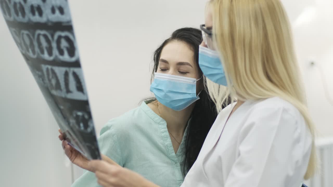 Woman doctor radiologist explains the results of CT scanning for young female patient, showing the snapshot with images, observing and analyzing CT scan in modern clinic beside modern CT scanner