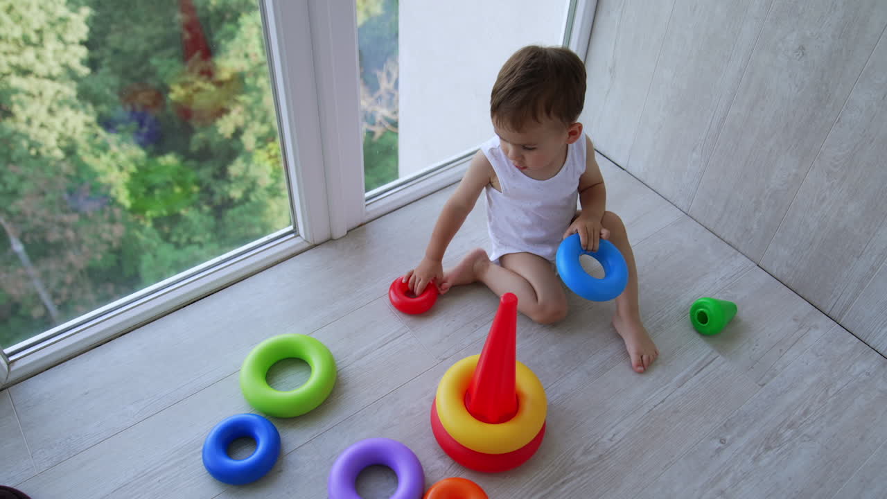 Tiny cute baby boy sitting on the floor at panoramic window. Focused toddler assembling the toy pyramid. Top view.