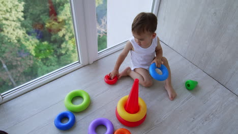 tiny cute baby boy sitting on the floor at panoramic window. focused toddler assembling the toy pyramid. top view.