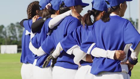 Happy-diverse-team-of-female-baseball-players-standing-on-field-with-arms-around-each-other