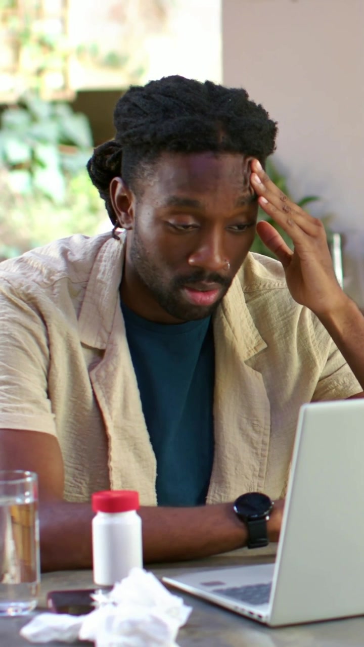 Vertical video: Showing fatigue man in teal at home holding head looking at laptop, meds, tissues