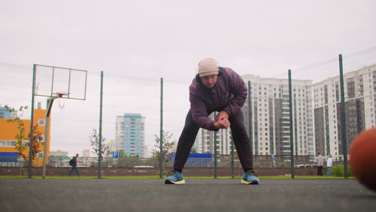 urban court white man dribbling basketball, overcast city setting, wearing hoodie and cap, focused on ball control and drills, low camera angle showing asphalt surface, fence and highrise buildings