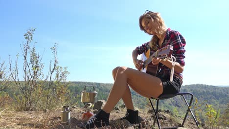 young blonde woman tourist  sitting on camping chair in nature and plays guitar