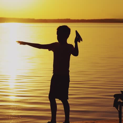 boy throws paper airplane. silhouette of a boy on an old bridge by the river. beautiful summer sunset