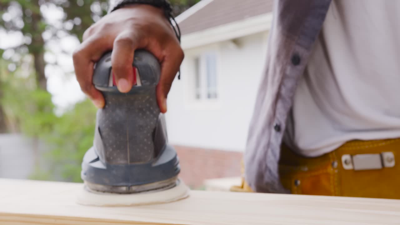 African American man holding orbital sander, sanding plank at home with yellow toolbelt to smooth