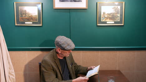 a mature man in a flat cap is sitting at a pub table, focused on reading a book. a glass on the table beside him, and a beige coat hangs on a nearby rack, creating a relaxed, contemplative atmosphere