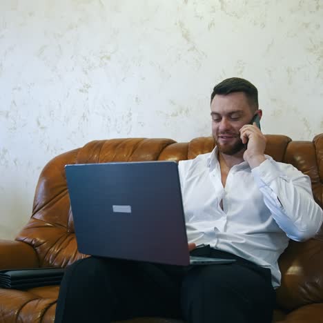 relaxed businessman is working in a hotel room on laptop. handsome man with laptop on knees speaks on the mobile phone