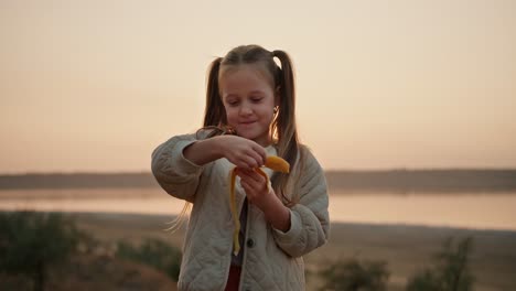 Portrait-of-a-little-blonde-girl-with-blue-eyes-in-a-white-jacket-who-peels-a-banana-and-eats-it-during-her-picnic-outside-the-city-in-the-evening-in-summer